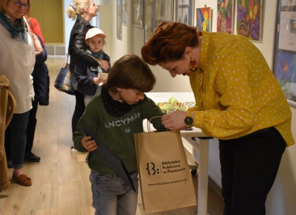 Artysta wraz z zastępcą dyrektora Sylwią Chojnacką-Tuzimek. Chłopiec patrzy co znajduje się w torbie z upomnikami.