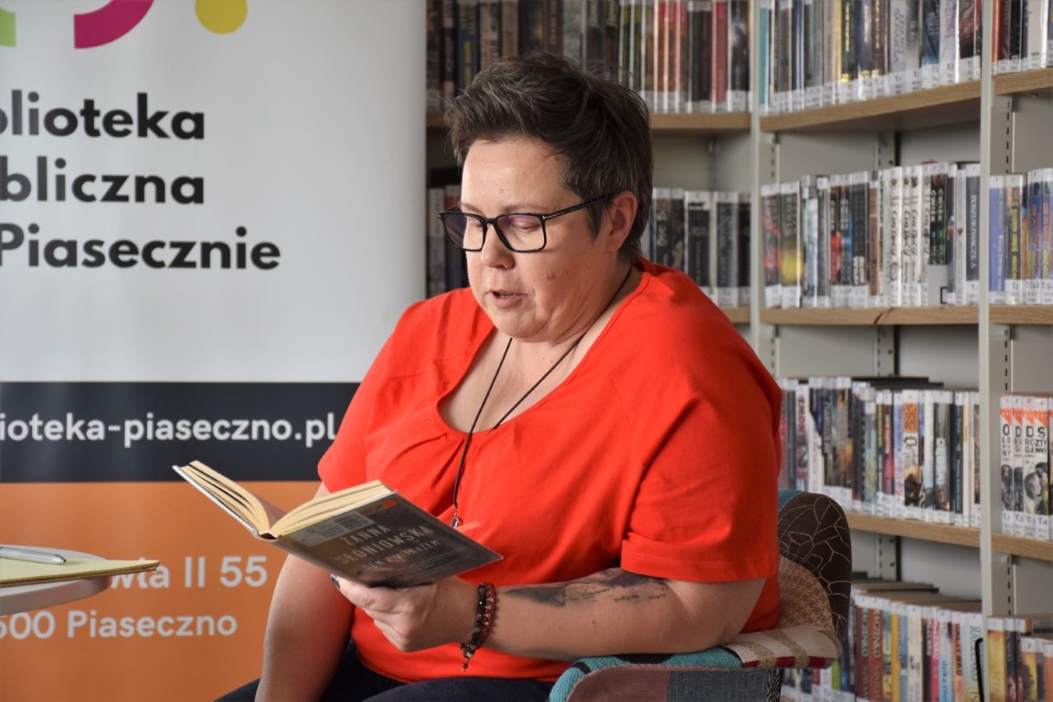 Prowadząca spotkanie Katarzyna Ostrowska na tle książek i baneru z logo Biblioteki Publicznej w Piasecznie cytuje fragment książki autorki.