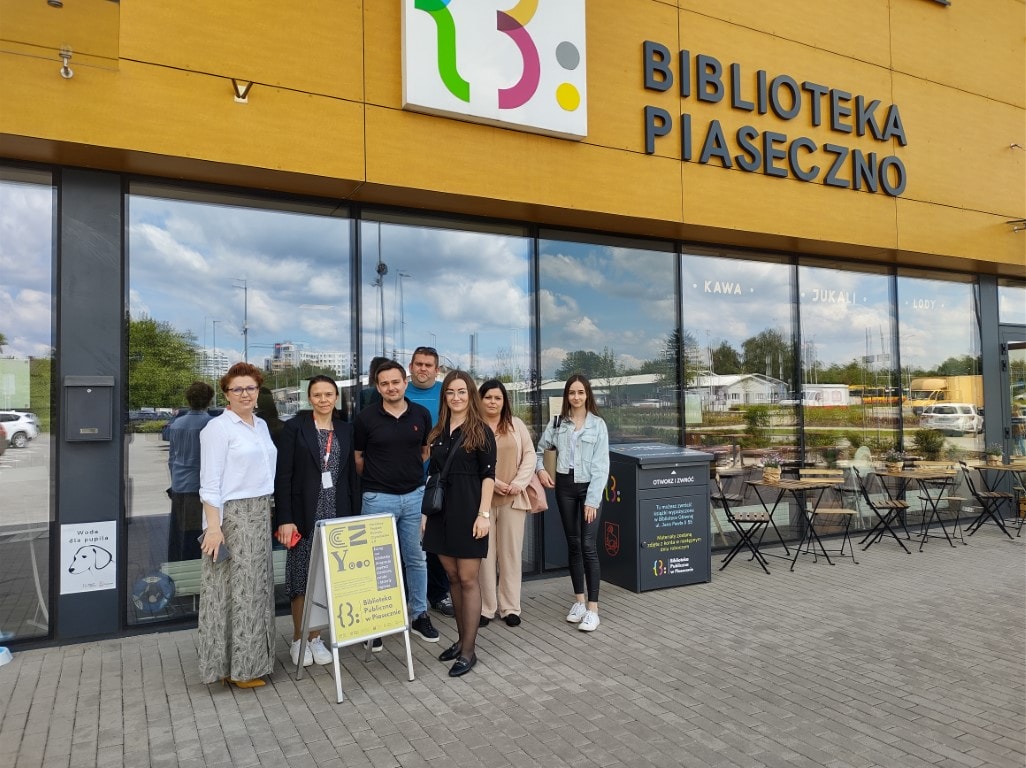Bibliotekarze z Białobrzegów wraz z zastępcą dyrektora Sylwią Chojnacką - Tuzimek oraz bibliotekarka Aldona Lazar pozują przed budynkiem biblioteki do zdjęcia i uśmiechają się.