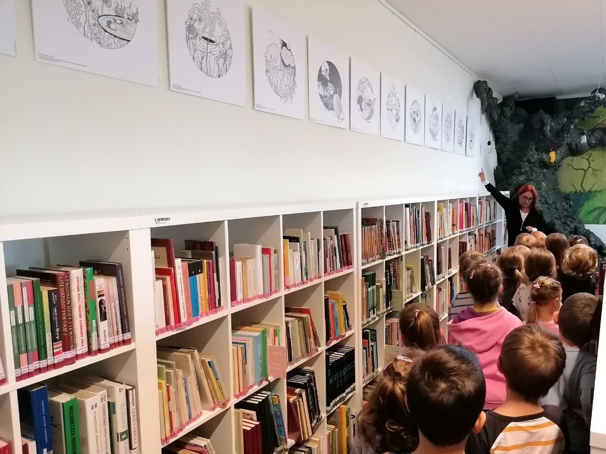 Pani bibliotekarka pokazuje dzieciom wystawę ilustracji do książki „Dzikie opowieści. Wędrówka tropem tajemnic natury”