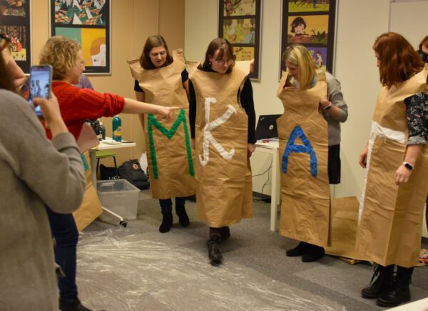 Uczestnicy warsztatów stoją ubrani w papierowe stroje z dużymi literami na środku. Od lewej widać następujące litery na strojach „M” „K” „A” „T”