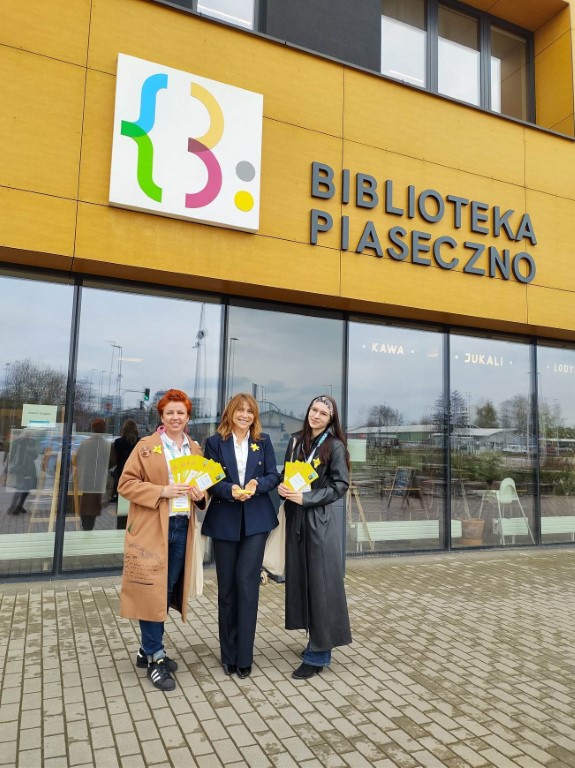 zastępca dyrektora Biblioteki Publicznej w Piasecznie oraz zastępca burmistrza razem z wolontariuszką-pracownicą biblioteki przed Biblioteką Główną w Piasecznie rozdają papierowe żonkile