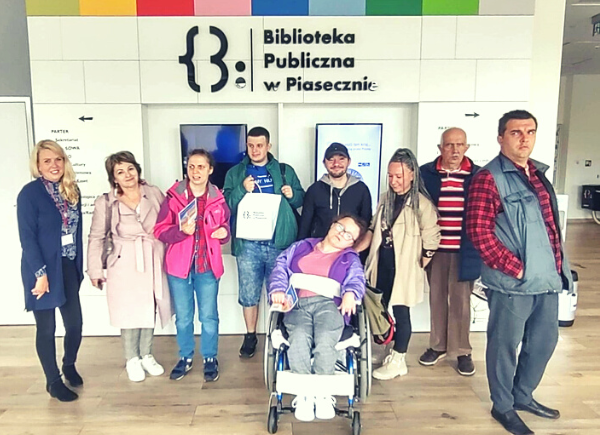 Wycieczka uczestników ŚDP W Pęcherach. Zdjęcie grupowe zrobione w korytarzy na parterze biblioteki z koordynatorką dostępności.
