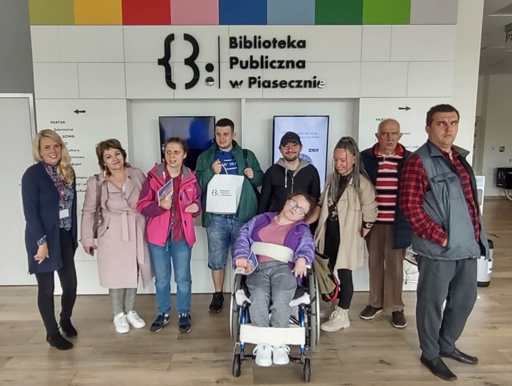 Wycieczka uczestników ŚDP W Pęcherach. Zdjęcie grupowe zrobione w korytarzy na parterze biblioteki z koordynatorką dostępności.