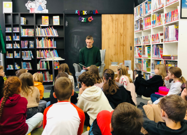 Autor Rafał Witek rozmawia z dziećmi siedzącymi na dywanie podczas spotkania autorskiego