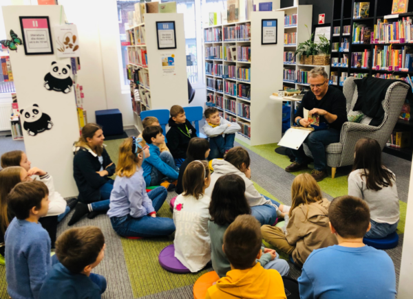 Autor Tomasz Trojanowski opowiada dzieciom siedzącym na dywanie o swoich ulubionych książkach z dzieciństwa