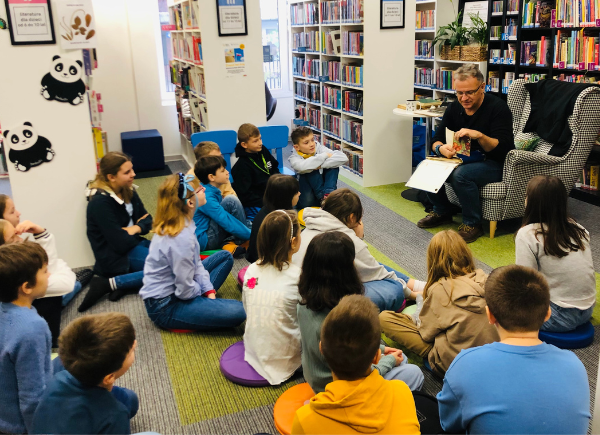 Autor Tomasz Trojanowski opowiada dzieciom siedzącym na dywanie o swoich ulubionych książkach z dzieciństwa