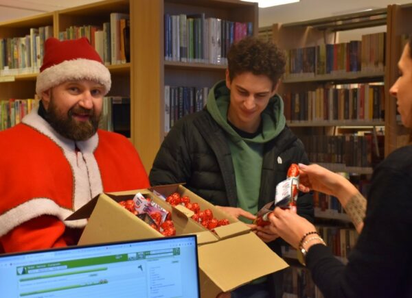 Jakub Ćwiek w stroju świętego mikołaja w Oddziale dla dorosłych w Bibliotece Publicznej w Piasecznie rozdający czekoladowe mikołaje po spotkaniu autorskim